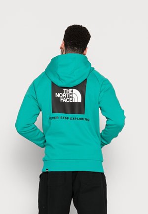 Mężczyzna ubrany w morski kaptur z logo The North Face i napisem „Never Stop Exploring” na plecach, stojący z rękami na biodrach.