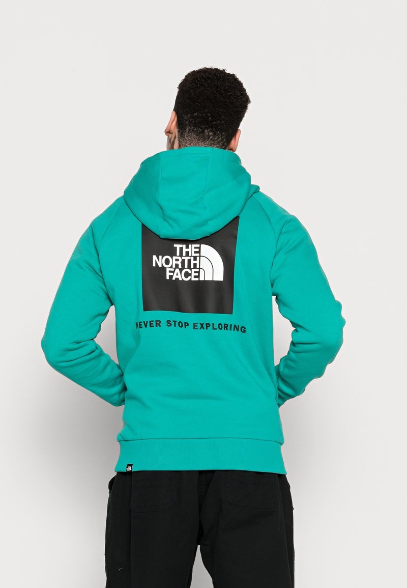 Homme portant un sweat à capuche bleu sarcelle avec le logo The North Face et l'inscription « Never Stop Exploring » au dos, debout les mains sur les hanches.