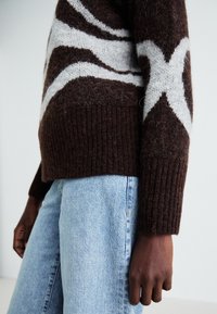 Brun strikket genser med hvite sikksakk-mønstre, med ribbet nederkant. Brukt med lyseblå denimjeans, som viser et avslappet, teksturert stoff.