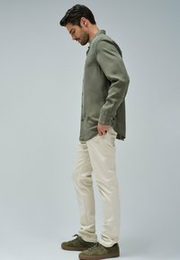 Olivno zelena srajca z gumbi, smetana hlače in olivno zeleni športni copati. Srajca ima sproščen kroj in dolge rokave, z gumbi spredaj.