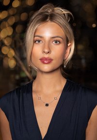 Zilveren ketting met een ronde zwarte hanger en twee bloemenaccenten; model draagt een marineblauwe jurk met geplooide textuur; zachte lichtachtergrond.