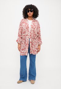 Červený a bílý květinový kimono kabát s páskem v pase, spárovaný s modrými rozšířenými džíny a béžovými botami. Model má na sobě černé sluneční brýle.