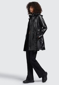 Cappotto impermeabile nero con finitura lucida, cappuccio e chiusure a bottoni. Abbinato a jeans neri svasati e sneakers nere.