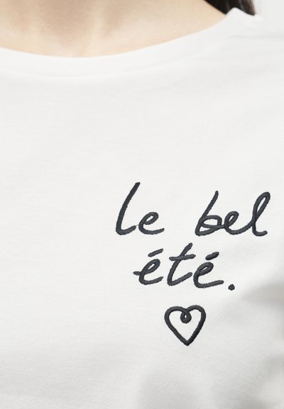 Λευκό πουκάμισο με μαύρο χειρόγραφο κείμενο "le bel été" και μια μικρή καρδιά από κάτω, φορεμένο από ένα άτομο που φαίνεται από το λαιμό και κάτω.