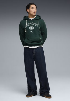 Jeune homme portant un sweat à capuche Manchester City vert foncé, un jean ample foncé et des baskets noires, debout les mains derrière le dos.