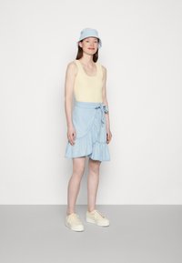Top de malha amarelo claro com bordas em formato de concha, combinado com uma saia envolvente azul com babados e uma cintura amarrada. Ténis bege.