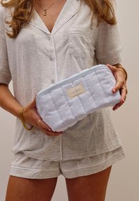 Lyseblå og hvitstripete kosmetikkveske med glidelås, polstret tekstur, og en etikett som leser "The Flat Lay Co." holdt av en person.