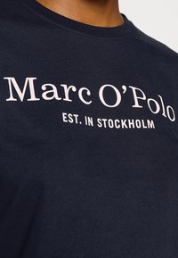 Námořnická modrá bavlněná trička s bílým potiskem "Marc O'Polo EST. IN STOCKHOLM." Měkká textura, kulatý výstřih a výrazný střih.