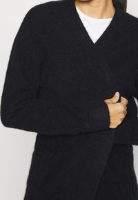 Cardigan negro y esponjoso con diseño de escote en V, mangas largas y cinturón en la cintura. El material parece suave y texturizado, ideal para superponer.