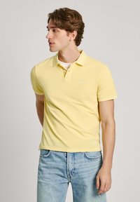 Žlté polo tričko so slim fit strihom, krátkymi rukávmi a golierom, spárované s svetlomodrými džínsami. Obsahuje vyšívané logo na hrudi.