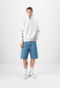 Sweatshirt cinza com capuz, calções de ganga azuis, meias brancas e sapatilhas brancas. Modelo está de pé contra um fundo branco liso.