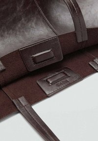 Binnenkant van een donkerbruine leren handtas met een gladde textuur, rechthoekige vakken en platte sluitingen. Opvallend geperst merklabel.