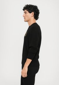 Μαύρο πουλόβερ με στρογγυλή λαιμόκοψη και ριμπ μανσέτες και στρίφωμα, με κυκλικό λογότυπο στο στήθος. Το ύφασμα φαίνεται λείο και εφαρμοστό.