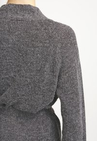 Tummanharmaa neulecardigan, jossa on teksturoitu pinta, huivikaulus ja sitomisnauha vyötäröllä; näkyvät saumat hihoissa.