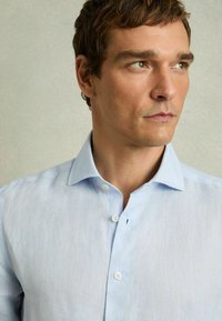 Camisa de lino azul claro con cuello, que presenta una textura sutil y cuatro botones en la parte frontal, exhibida sobre un fondo neutro.