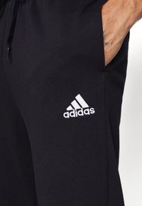 Čierne športové nohavice z mäkkého materiálu, s elastickým pásom, bočnými vreckami a bielym logom Adidas na ľavom stehne.