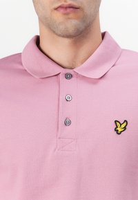 Lys rosa polo skjorte laget av teksturert stoff med nedbrettbar krage og tre knapper. Har en svart og gul brodert logo.