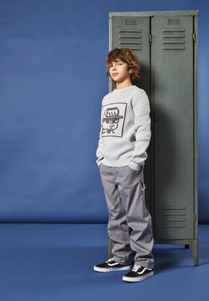 Suéter gris claro con diseño gráfico, pantalones cargo gris claro, zapatillas negras, de pie junto a un casillero verde contra un fondo azul.