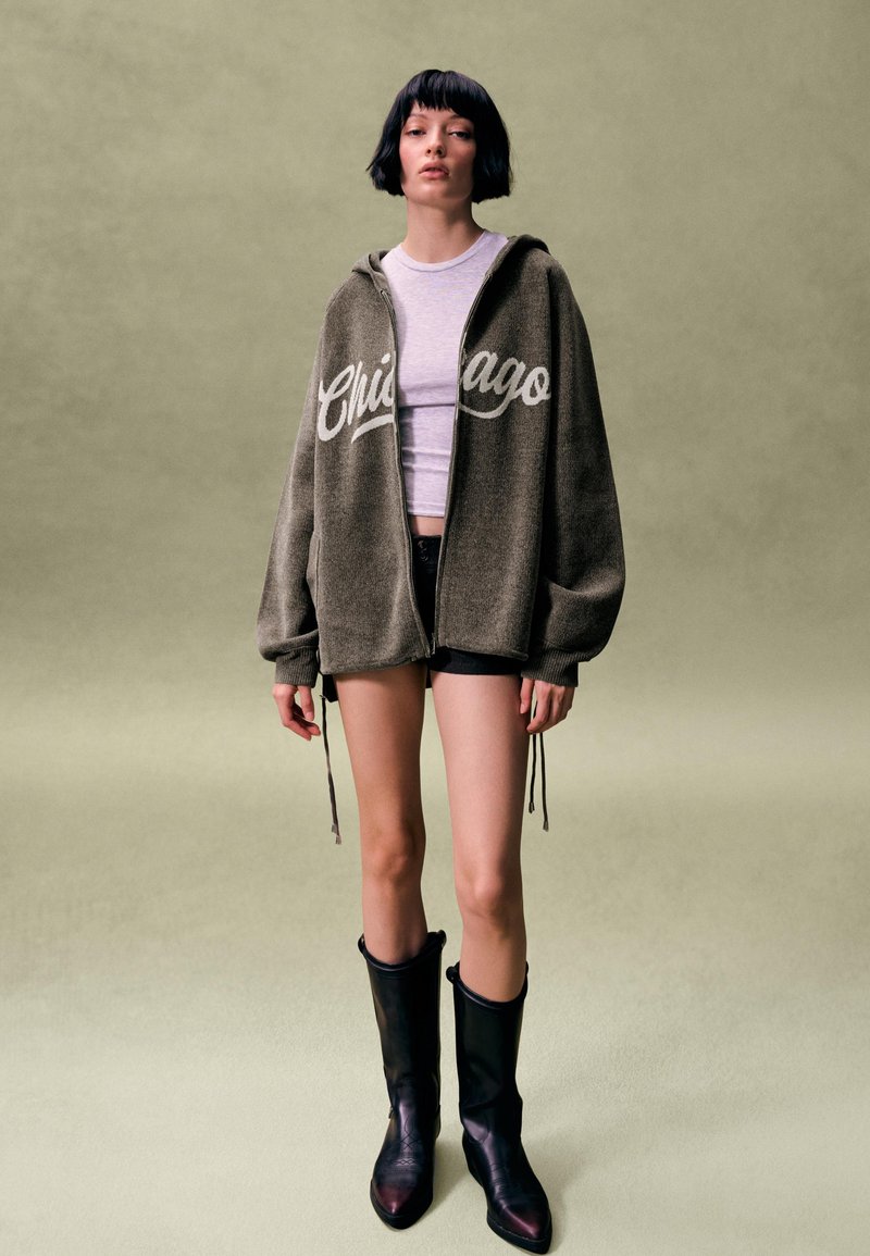 Žena s krátkymi čiernymi vlasmi má na sebe veľkú sivú mikinu na zips s nápisom „Chicago“, biely krátky top, čierne krátke nohavice a čierne čižmy po stred lýtok.