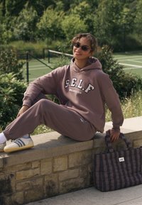 Helepruun kapuutsiga dressipluus tekstiga "S.E.L.F.", sobivad dressipüksid ja valgete triipudega tossud. Lähedal ruuduline lilla käekott käepidemetega.