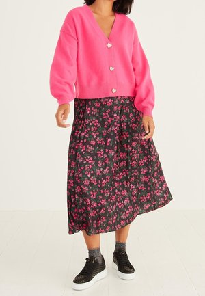 Vrouw draagt een felroze vest met knopen in de vorm van hartjes over een zwarte midirok met roze bloemenprint en zwarte sneakers met grijze sokken.