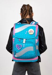 Ein türkisfarbener Rucksack mit pinken Akzenten, der mit Punkten, einem silbernen Wellenmuster und dekorativen Pins, darunter ein Flamingo und ein Herzchen, verziert ist.