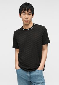 Μαύρο μπλουζάκι με ολόκληρο διακριτικό μοτίβο λογότυπου MK, στρογγυλή λαιμόκοψη με λευκή διακόσμηση. Συνδυάζεται με ανοιχτόχρωμο τζιν τζιν.