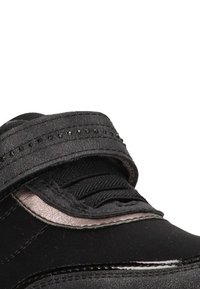 Deportiva negra con cordones negros y una correa negra texturizada que presenta pequeños agujeros decorativos a lo largo de la parte superior.