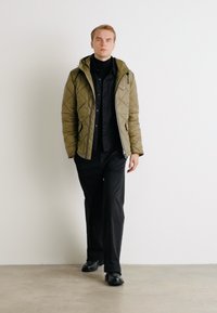 Veste matelassée vert olive avec capuche, portée sur un col roulé noir. Pantalons et chaussures noirs complètent la tenue. Design simple et structuré.