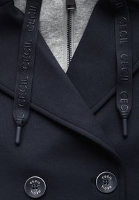 Gros plan d'un manteau bleu marine avec des boutons noirs et des cordons étiquetés "CECIL", superposé à un vêtement gris à fermeture éclair.