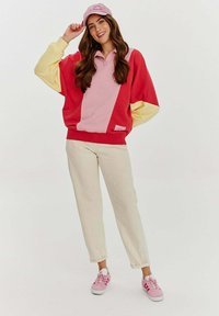 Barevná oversized mikina v červené, růžové a žluté barvě se sportovním designem. V kombinaci se světle béžovými kalhotami a růžovými teniskami.