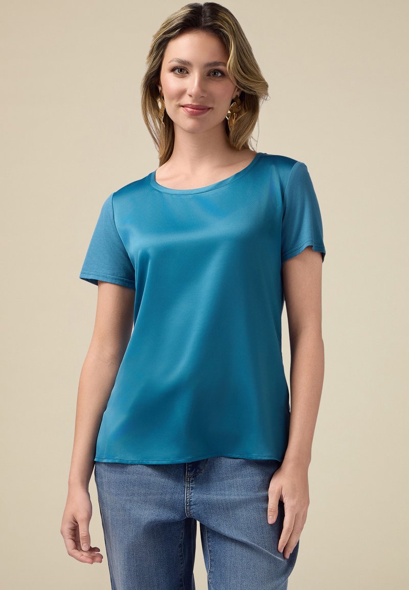 Top scurt de culoare teal, dintr-un material neted, cu guler rotund și o croială lejeră, asortat cu blugi din denim albastri.