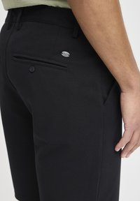 Shorts noirs en mélange de coton, avec une poche arrière à bouton et une étiquette de marque en métal. Des poches latérales sont également visibles.