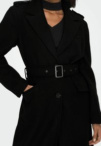 Manteau en mélange de laine noire avec col à revers, taille ceinturée, fermeture à boutons et poches latérales. Présente une silhouette ajustée et une texture lisse.