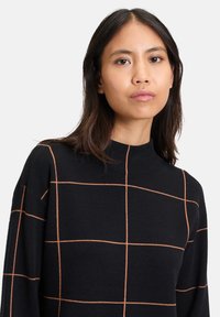 Svart tröja med orange rutmönster, hög hals och långa ärmar. Mjuk textur, avslappnad passform och synliga sömdetaljer.