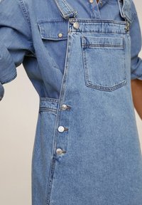 Vaateyliopistot vaaleansinisestä denimiä säädettävillä olkaimilla ja metalliosilla. Siinä on rintatasku ja viiden napin sivukiinnitys.