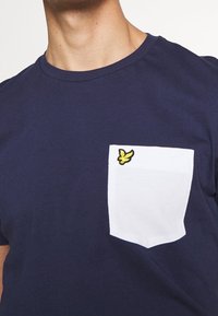 Námořnické bavlněné tričko s bílou náprsní kapsou a žlutě vyšitým logem ptáka. Krátké rukávy a kulatý výstřih.