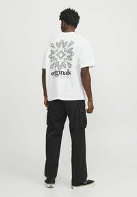 T-shirt bianca in cotone con una grande stampa geometrica nera e il testo "originals STUDIO" sulla parte posteriore, abbinata a pantaloni cargo neri.