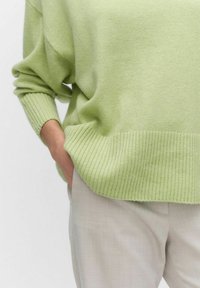 Jasnozielony, oversize sweter z prążkowanymi mankietami i dołem, wykonany z miękkiego materiału. Połączony z jasnymi, teksturowanymi spodniami, ręka w kieszeni.