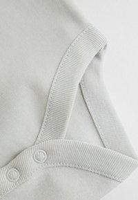 Šviesiai pilkas medvilninis bodis su gofruotais kraštais ir spaustukais. Turi lygią tekstūrą ir be siūlių siuvimą ties kaklo ir pažastų siūlėmis.