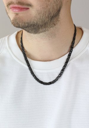 Black chain necklace with a textured pattern, featuring interlocking links. Worn over a white textured shirt.