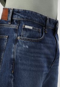 Dark blue denim jeans with a light brown leather patch, front pockets, and contrast stitching. Features a faded accent on the fabric.