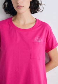 Lys rosa T-skjorte med korte ermer og rund hals, med lomme og hvit blomsterutforming på lommen. Mykt bomullsmateriale.