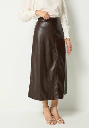 Vrouw die een hooggetailleerde donkerbruine leren midirok draagt met een witte blouse met lange mouwen en gouden open-teenhakken.