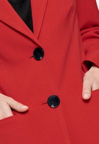Blazer rojo hecho de tela texturizada con solapas. Presenta dos botones negros y bolsillos laterales.