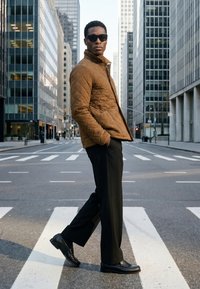 Hombre con chaqueta marrón acolchada, pantalones negros y gafas de sol cruzando un paso de peatones en una calle de la ciudad entre altos edificios de oficinas.