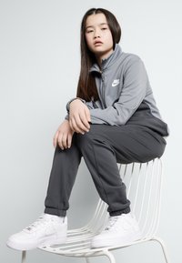 Ensemble de survêtement gris Nike avec une veste zippée et un pantalon assorti, arborant un logo blanc. Des baskets blanches complètent l'ensemble.