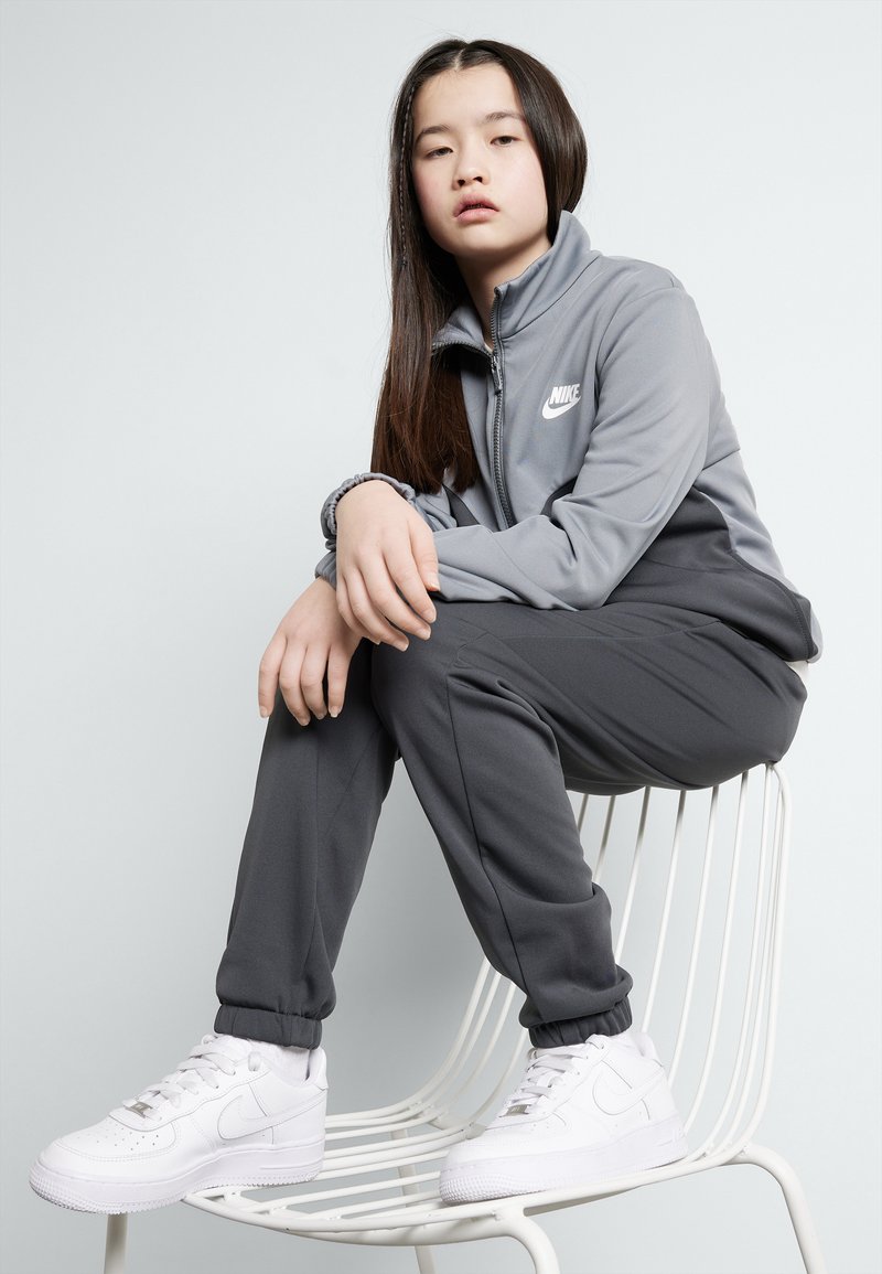 Ensemble de survêtement gris Nike avec une veste zippée et un pantalon assorti, arborant un logo blanc. Des baskets blanches complètent l'ensemble.