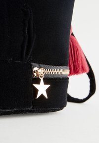 Schwarze Veloursledertasche mit Reißverschluss, versehen mit einem metallischen Sternanhänger und einem rosa Quastenakzent an der Seite. Flache, rechteckige Form.