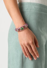 Pink translucent bangle featuring green accents and ornamental details, worn on a hand. The textured background is soft green fabric.